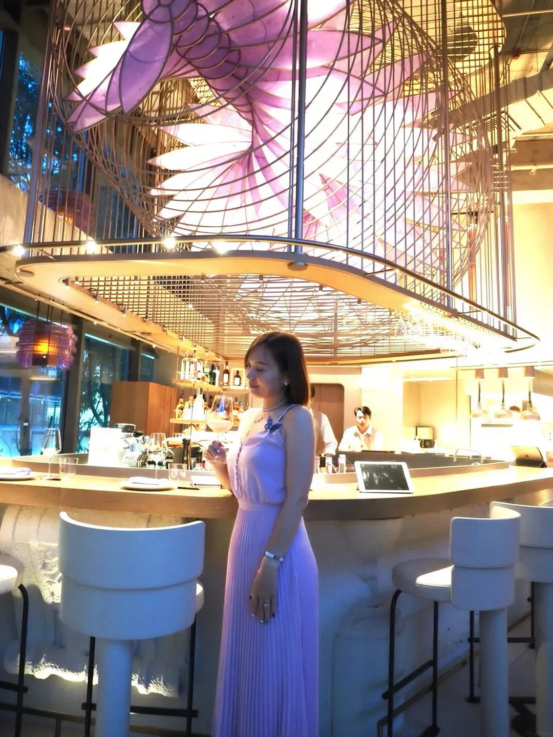 Alicia in a violet pleated dress beneath a sculptural light installation
